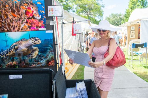 A woman in a large hat examines artwork at an outdoor art festival, surrounded by colorful displays and attendees.