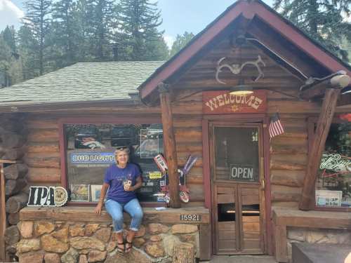 A woman sits on the stone steps of a rustic log cabin with a welcoming sign, surrounded by trees and outdoor decor.