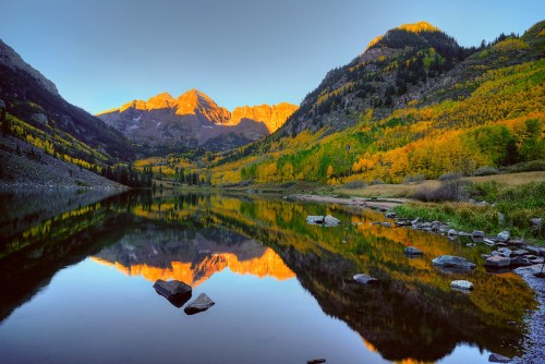 A serene lake reflects golden autumn trees and mountains at sunrise, creating a picturesque landscape.