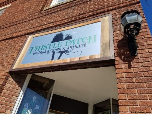 Sign for "Thistle Patch Vintage Garden & Antiques" on a brick building, featuring decorative elements and a lantern.