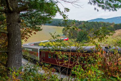 A vintage train travels through a scenic landscape, surrounded by trees and rolling hills in autumn colors.