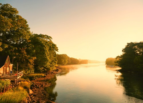 A serene river scene at sunrise, with lush trees and a small wooden structure along the water's edge.