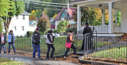 A group of people walking past a house with a porch and a well-maintained garden on a sunny autumn day.