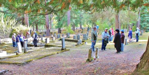 A group of people gathers in a serene cemetery surrounded by trees, engaging in conversation among the gravestones.