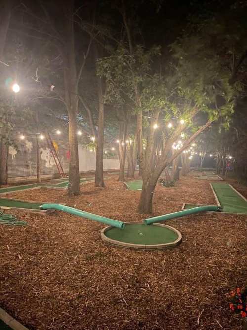 A dimly lit mini-golf course surrounded by trees, with green turf and string lights creating a cozy atmosphere.