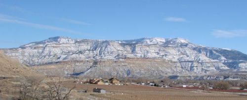 Snow-covered mountains rise above a valley with scattered houses and fields under a clear blue sky.