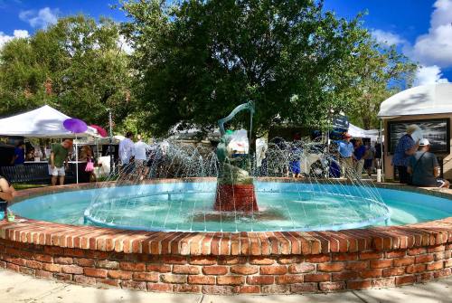 A fountain with a statue in the center, surrounded by people and tents at an outdoor event on a sunny day.