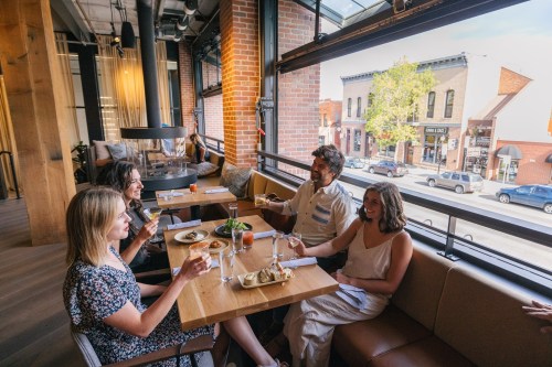 Four friends enjoy drinks and food at a cozy restaurant with large windows overlooking a street.