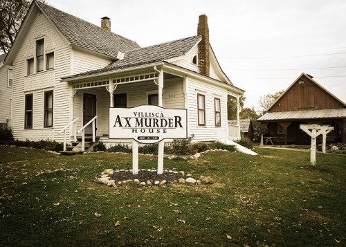 Historic Villisca Ax Murder House with a sign in front, surrounded by grass and trees, under a cloudy sky.