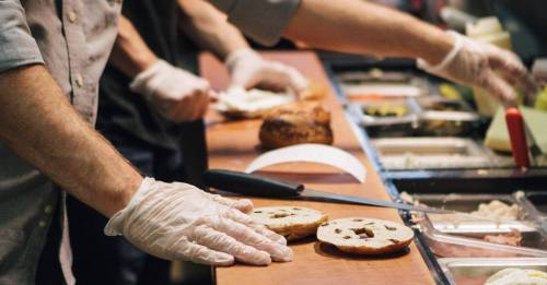A person in gloves prepares a sandwich with various toppings on a counter filled with ingredients.