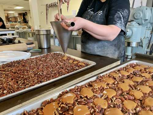 A person pours caramel into a tray of pecans, preparing a dessert in a kitchen setting.