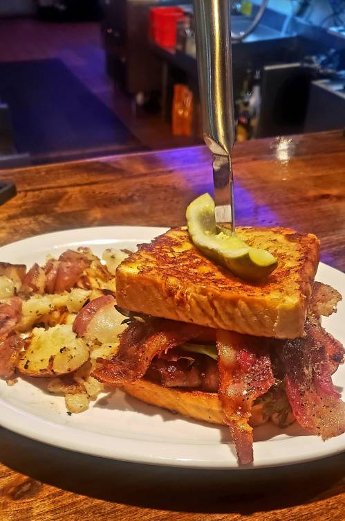 A hearty sandwich with bacon and pickles, served with a side of crispy potatoes on a white plate.
