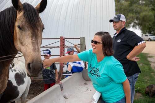 A woman in a teal shirt smiles as she reaches out to a horse, while a man stands nearby and children play in the background.
