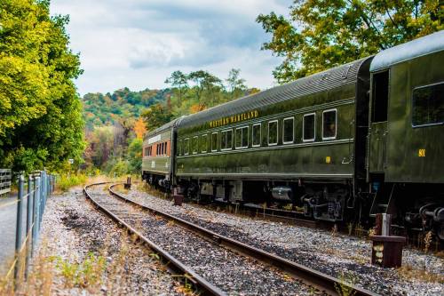A vintage train car parked beside a scenic railway surrounded by trees and autumn foliage.