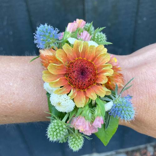 A colorful floral bracelet made of various flowers and greenery, worn on a wrist against a dark background.