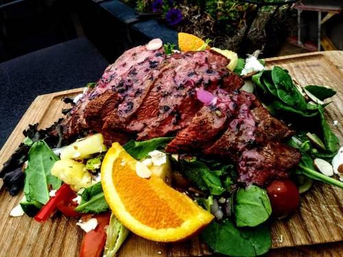 A beautifully plated steak on a bed of fresh greens, cherry tomatoes, and orange slices, garnished with herbs.
