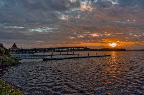 Sunset over a calm lake with a pier and bridge in the background, surrounded by clouds and a hint of orange sky.