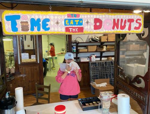 A person in a pink sweatshirt and cap holds a donut menu at a shop with a colorful "Time to Eat the Donuts" sign.