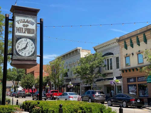 A clock tower sign reads "Village of Milford" with shops and cars lining a sunny street under blue skies.