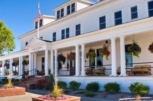 Historic hotel exterior with white columns, hanging flower baskets, and a welcoming porch on a sunny day.