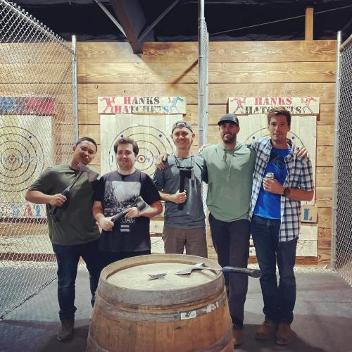 Five people stand in front of wooden target boards, holding axes and smiling, with a barrel in the foreground.