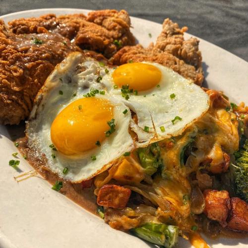 A plate of fried chicken with two sunny-side-up eggs, cheese, and sautéed vegetables, garnished with chives.
