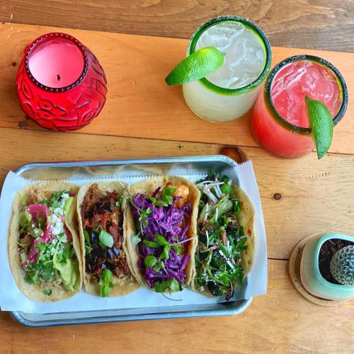 A tray of four colorful tacos with various toppings, alongside two cocktails garnished with lime, on a wooden table.