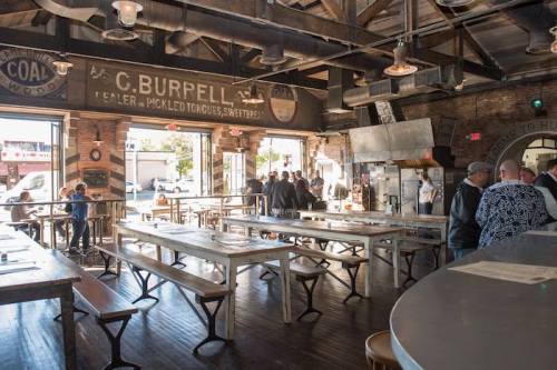 Interior of a rustic restaurant with wooden tables, exposed beams, and patrons mingling near a large counter.