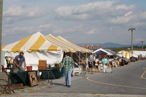 A bustling outdoor market with yellow-striped tents, vendors, and shoppers browsing antiques and collectibles.