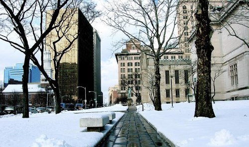 A snowy cityscape featuring a pathway lined with trees and buildings, including a tall glass structure in the background.