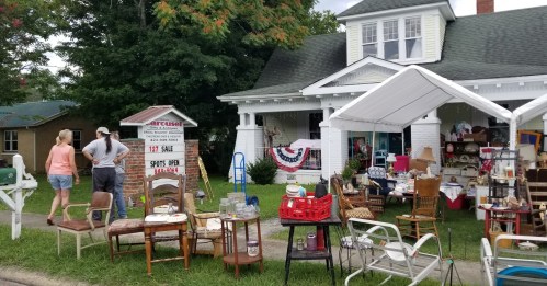 A yard sale in front of a white house, featuring various items for sale under a tent, with two people browsing.