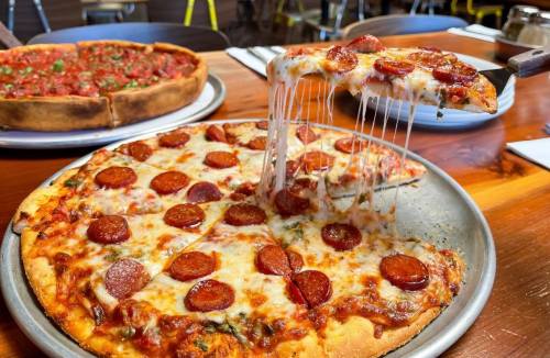 A close-up of a pepperoni pizza with melted cheese being lifted from a metal tray, with another pizza in the background.