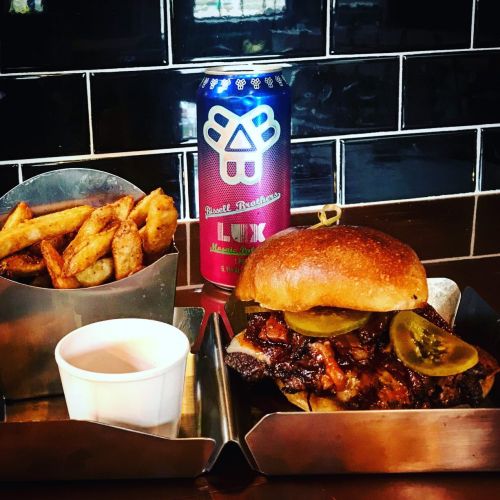 A delicious burger with pickles, served with fries and a can of beer on a wooden table.