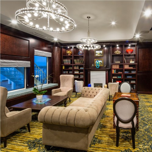 A stylish room with a cozy sofa, elegant chairs, a bookshelf, and modern chandeliers, featuring large windows and a warm ambiance.