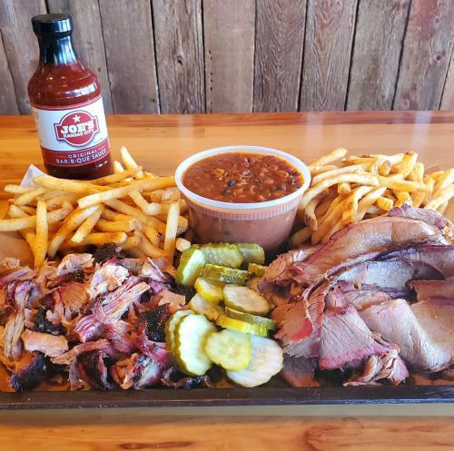 A platter of barbecue meats, pickles, fries, and baked beans, with a bottle of barbecue sauce in the background.