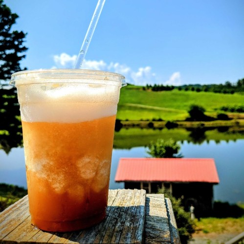 A refreshing drink in a clear cup sits on a wooden surface, with a scenic view of a lake and green hills in the background.
