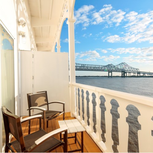 A balcony with two chairs and a table overlooks a river and a bridge under a blue sky with scattered clouds.