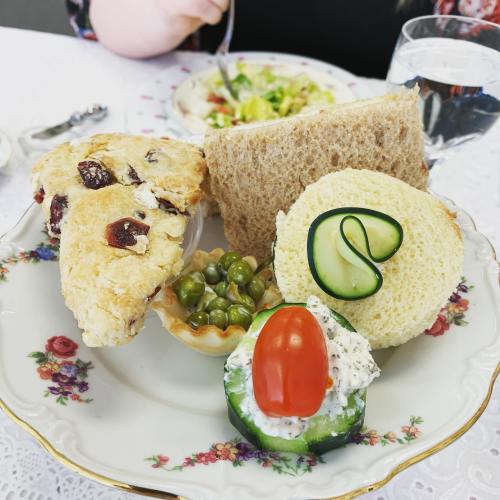 A plate with a scone, sandwich, cucumber salad, and a small dish of peas, set on a floral-patterned tablecloth.