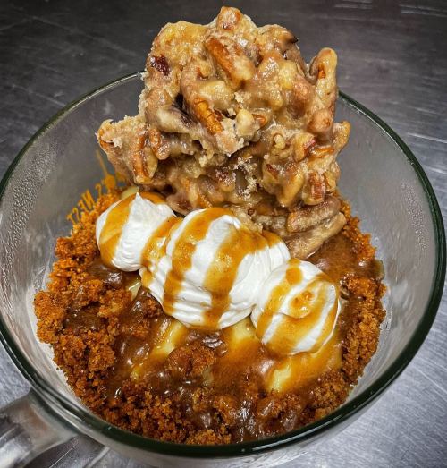 A glass dish filled with crumbled cookie, caramel sauce, whipped cream, and a pecan topping.