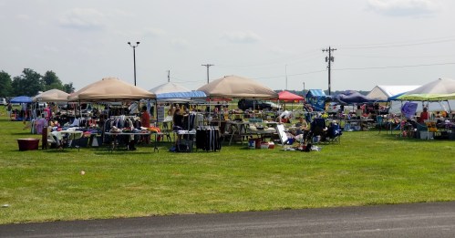 A bustling outdoor market with various tents and tables displaying clothes and goods on a grassy field.