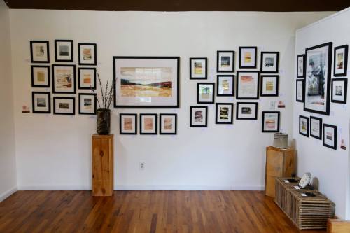 A gallery wall featuring various framed artworks and photographs, with wooden display stands and a warm wooden floor.