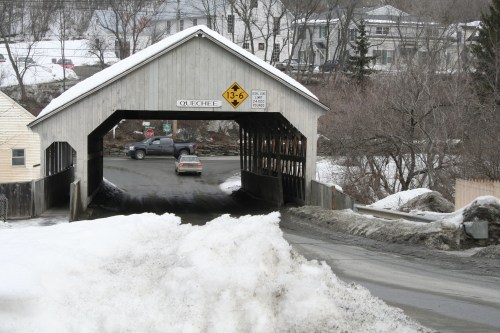 A snow-covered road leads to a wooden covered bridge with a vehicle approaching, surrounded by winter scenery.