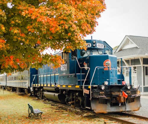A blue train engine parked near a station, surrounded by vibrant autumn foliage.