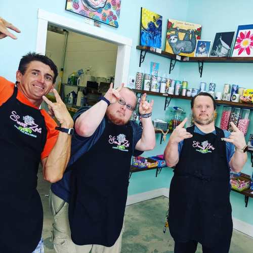 Three people in aprons pose playfully in a colorful art studio filled with supplies and artwork on the walls.