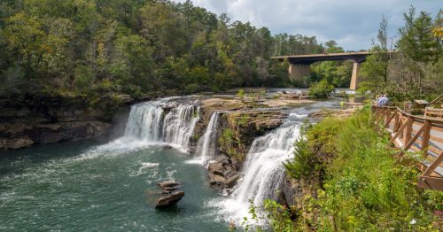 A scenic view of a waterfall cascading into a river, surrounded by lush greenery and a bridge in the background.