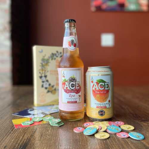 Two bottles of ACE hard cider (Rose and Mango) on a wooden table, surrounded by colorful game tokens and a decorative box.