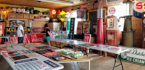 A vintage garage filled with retro signs, tables displaying memorabilia, and people browsing the items.