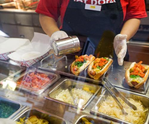 A vendor prepares hot dogs, adding toppings like diced tomatoes and onions from a display of fresh ingredients.
