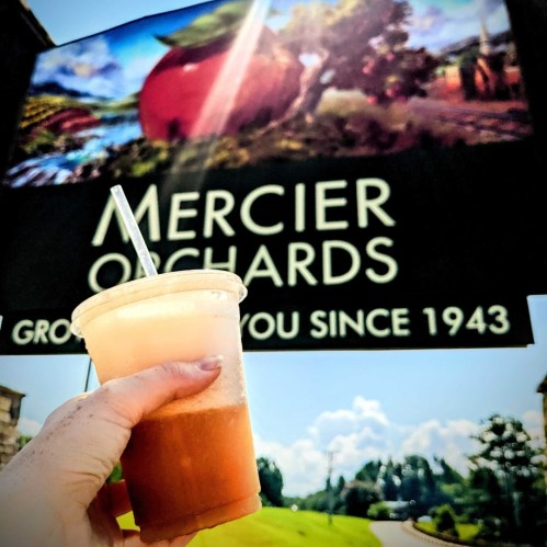 A hand holding a drink in front of a sign for Mercier Orchards, with blue skies and clouds in the background.