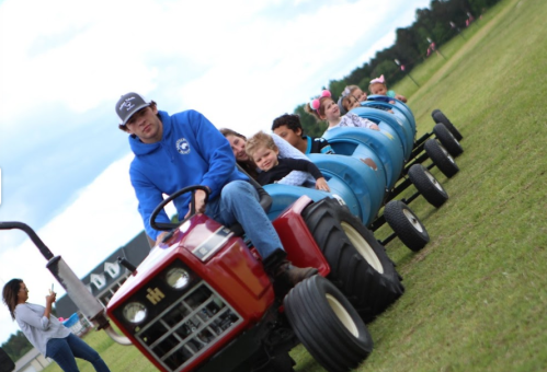 A man drives a small tractor pulling a blue barrel train with children enjoying a ride on a grassy field.